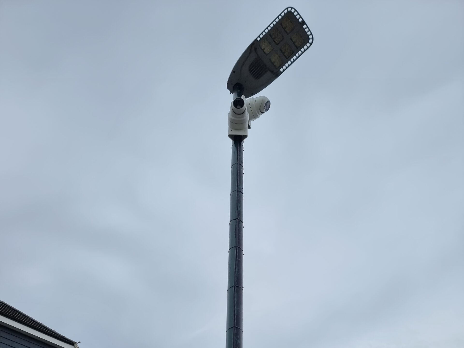 A high‑mounted CCTV camera installed beneath an LED floodlight on a tall pole to monitor an exterior site boundary.