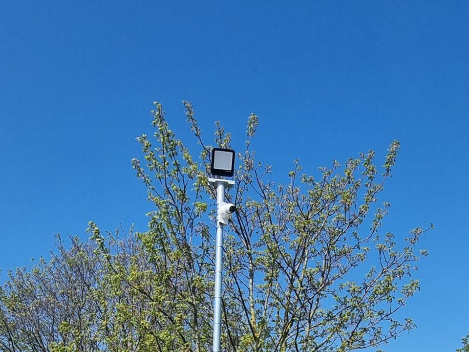 CCTV camera mounted on a security pole monitoring the parking area and boundary fence of a commercial storage yard.