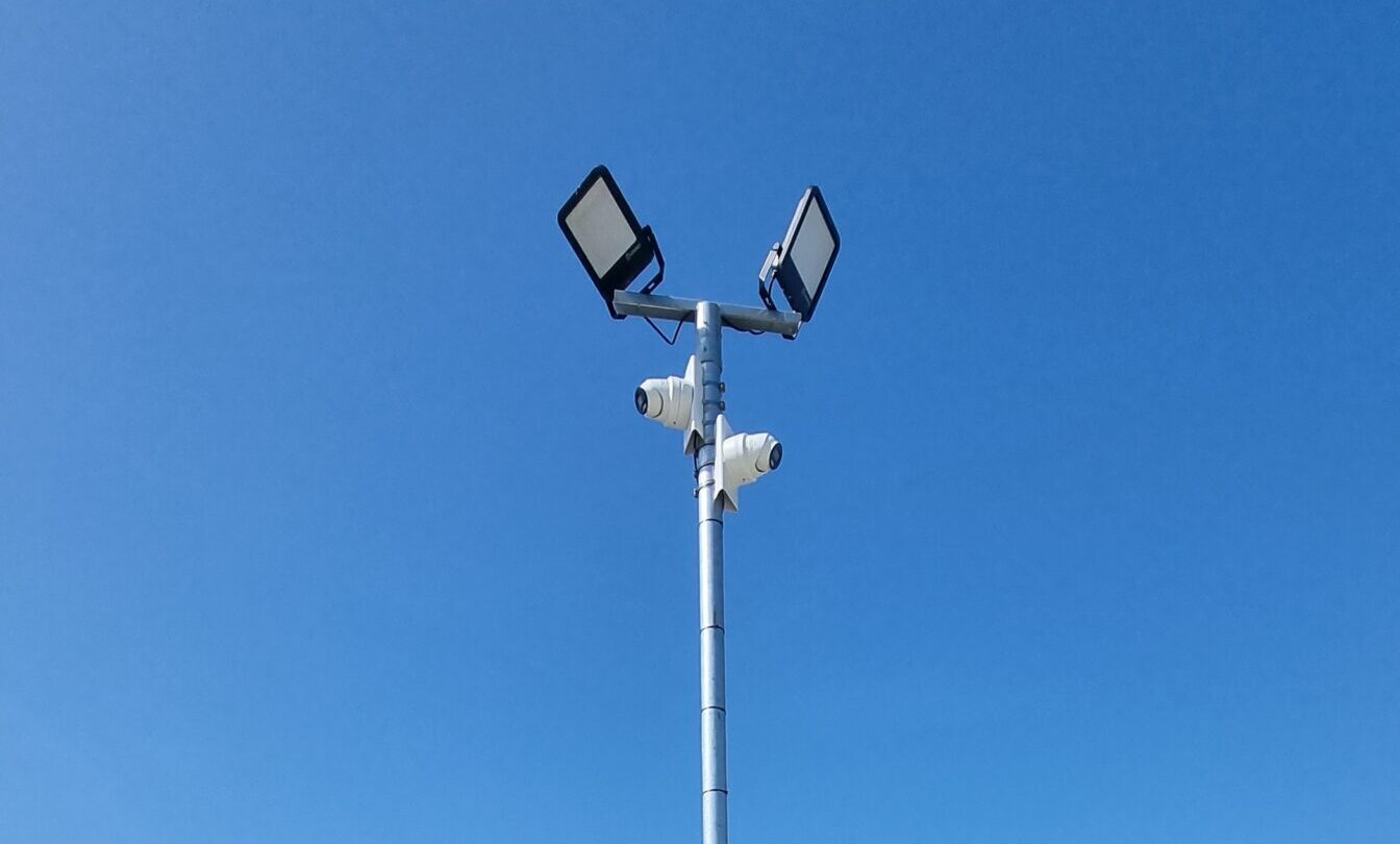 CCTV cameras installed on a pole overlooking the entrance and perimeter fencing of a commercial storage yard.