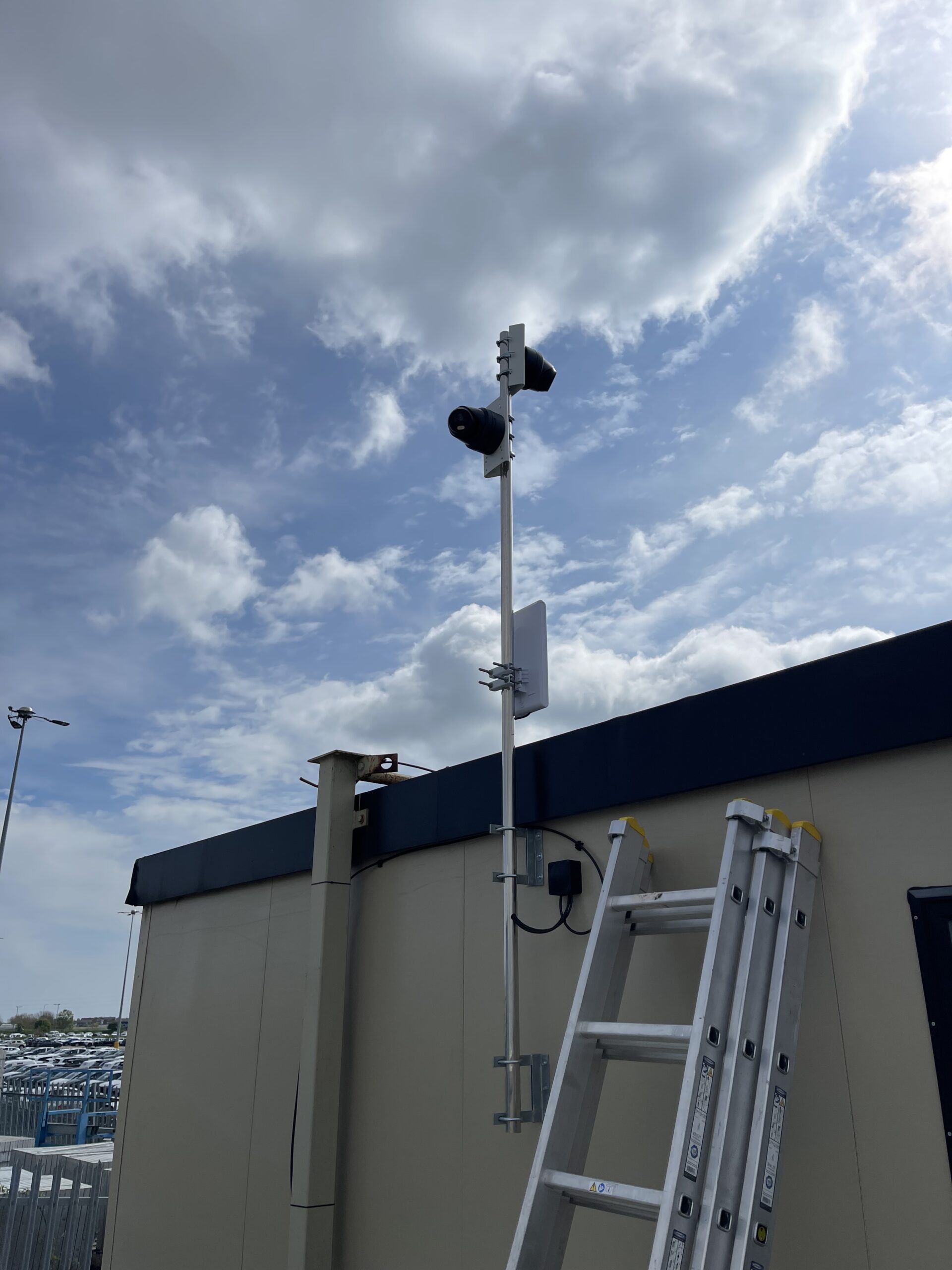 Two CCTV cameras mounted high on a pole fixed to a cabin building, providing perimeter surveillance for a commercial site.