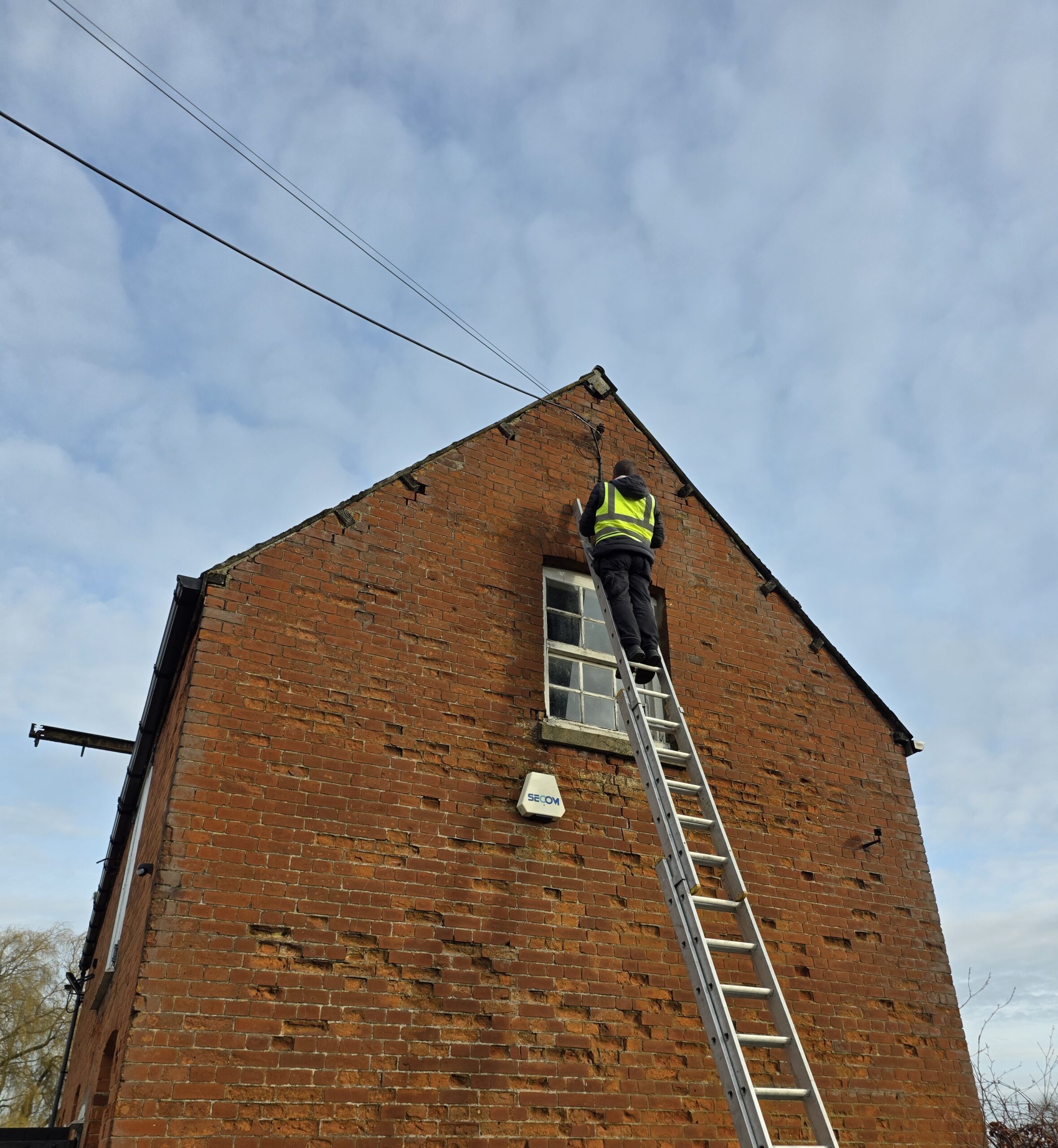 An engineer on a ladder installing cable to the exterior of a brick building as part of a CCTV or security system setup.