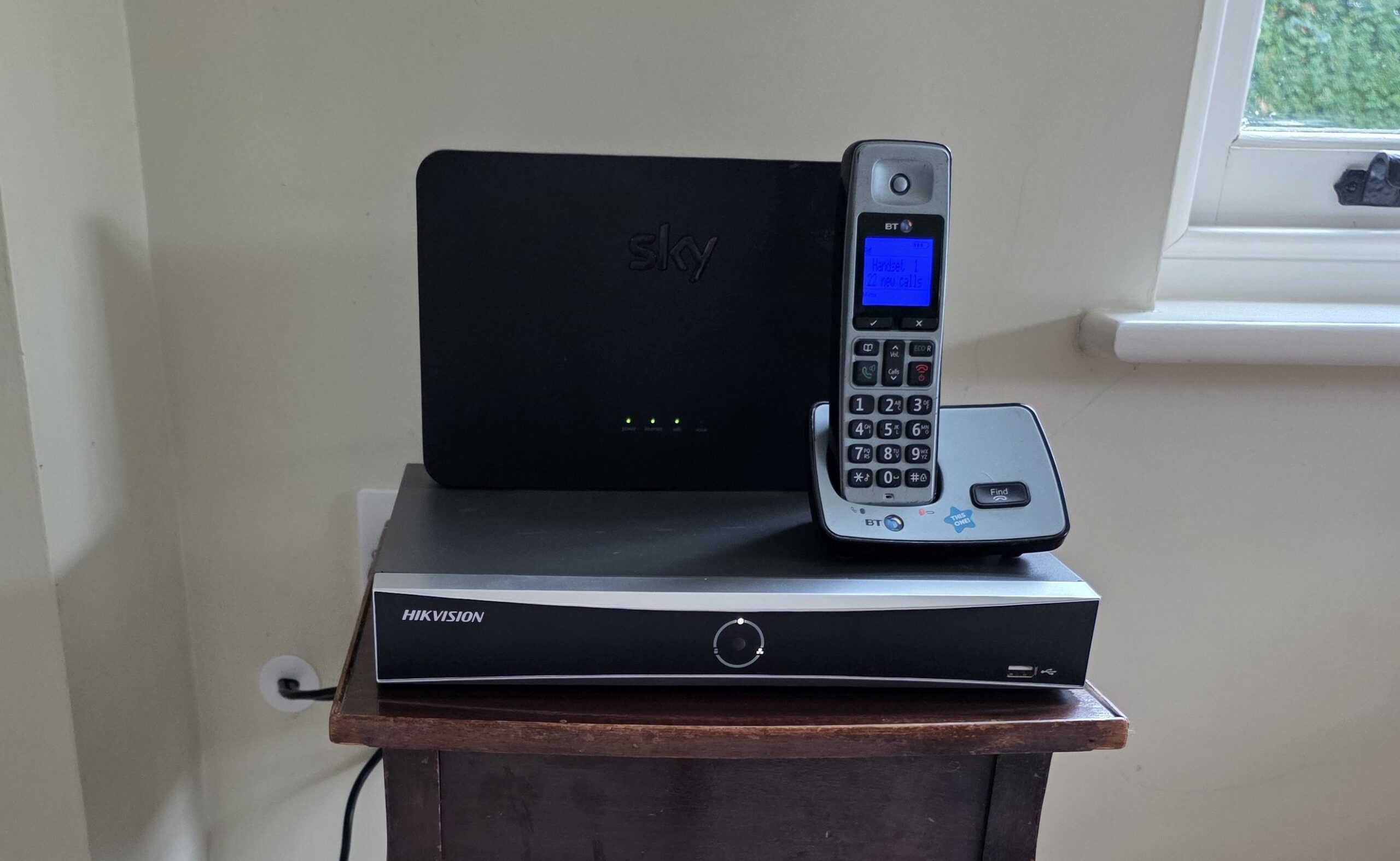 A Hikvision CCTV recorder placed beneath a broadband router and cordless phone as part of a home security system installation.