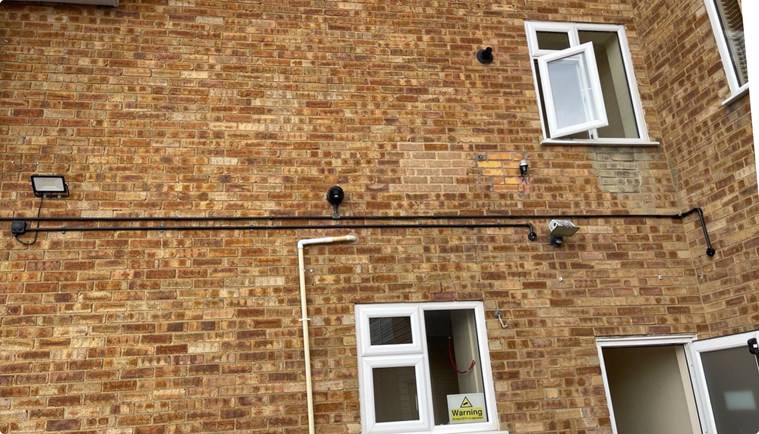 CCTV camera and external cable containment installed along the brick exterior wall of a commercial building to improve property security.