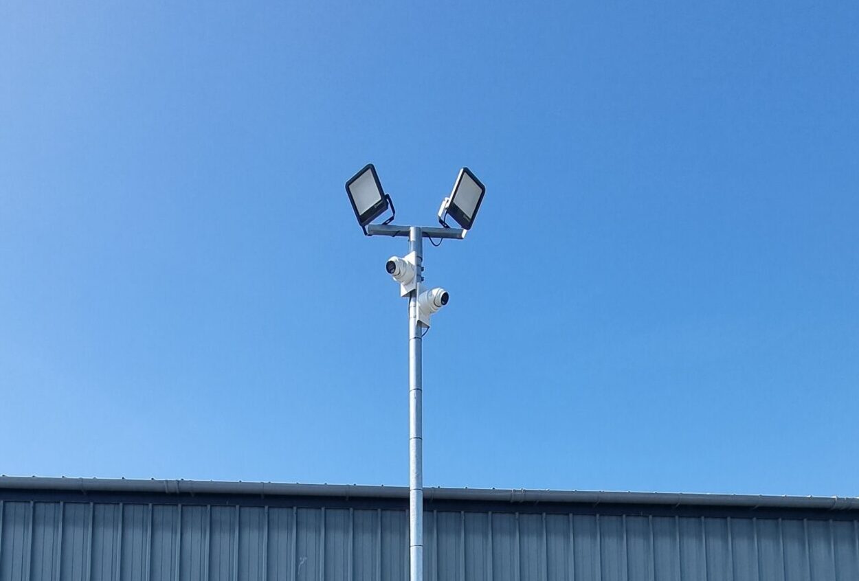CCTV and floodlight pole positioned along warehouse fencing to enhance perimeter security at a commercial storage yard.